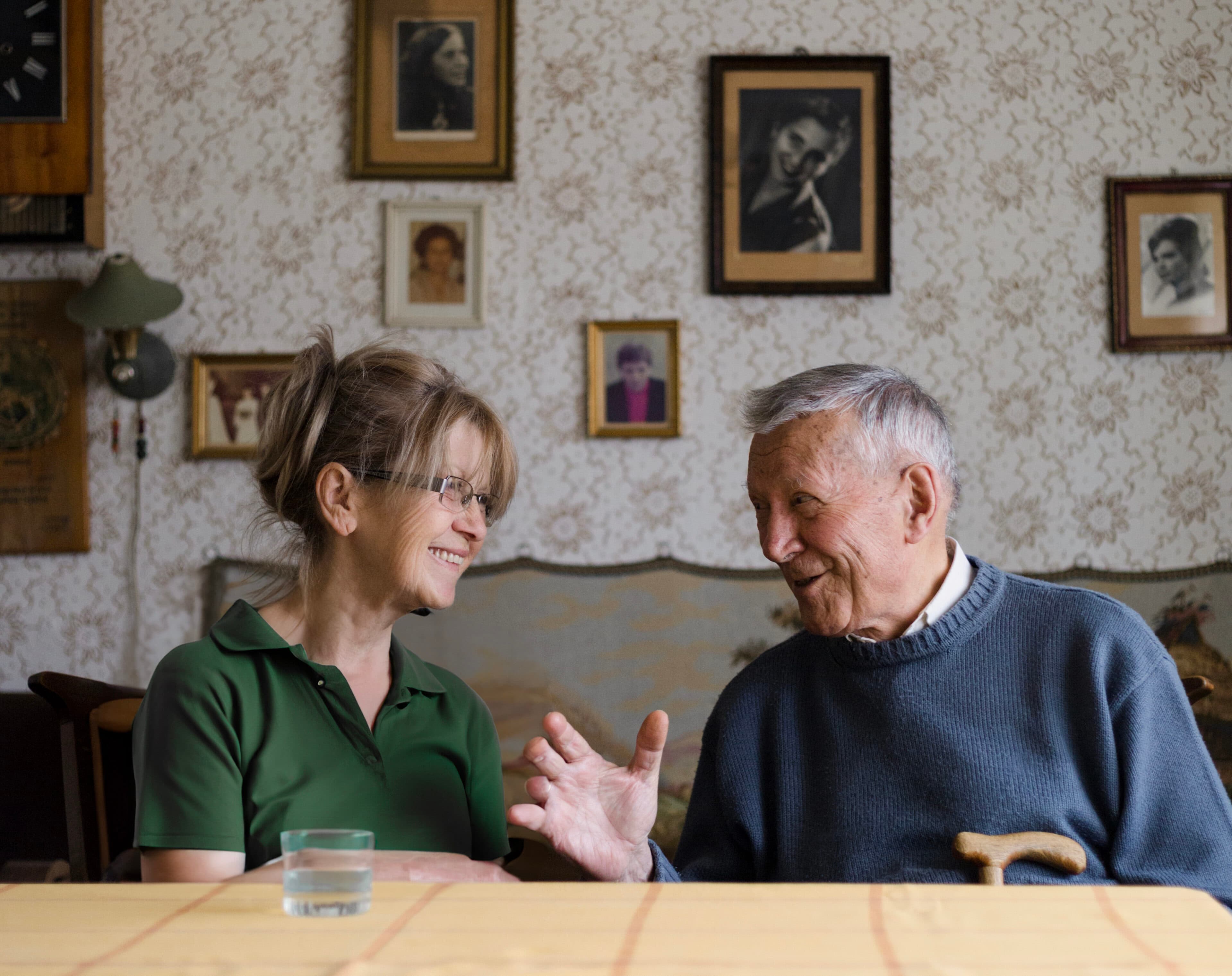 An elderly man and a woman smile and talk at a table, with framed photos on the wall behind them. - Home Instead