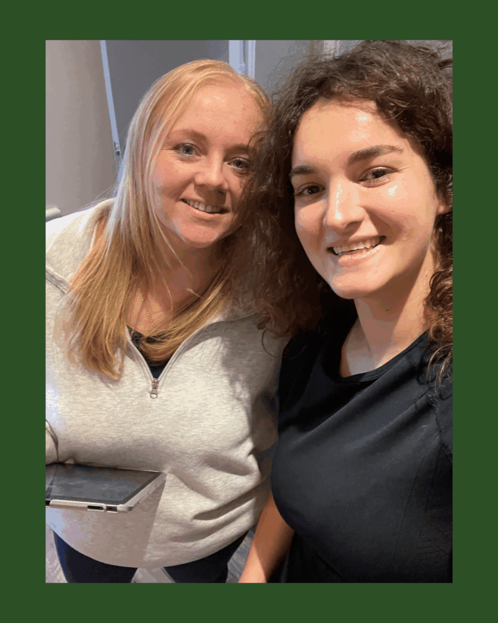 Two women smiling for a selfie indoors, one holding a laptop; green border round the photo. - Home Instead