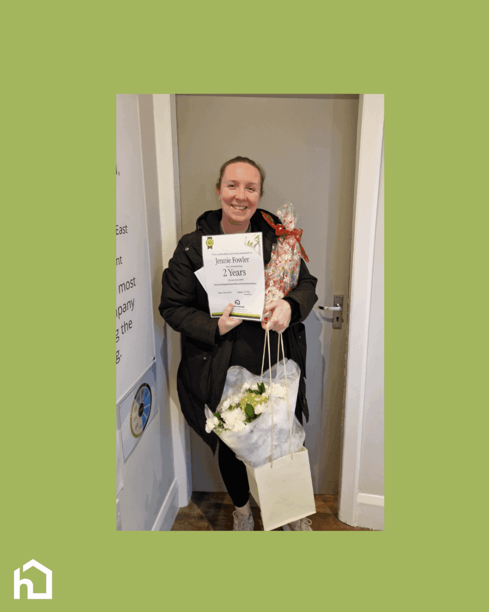 Woman smiling, holding a 2 Years award certificate, flowers, and a wrapped present, standing by a door. - Home Instead