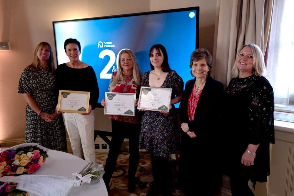 Six women stand indoors, three holding certificates, in front of a screen displaying the Home Instead logo. - Home Instead