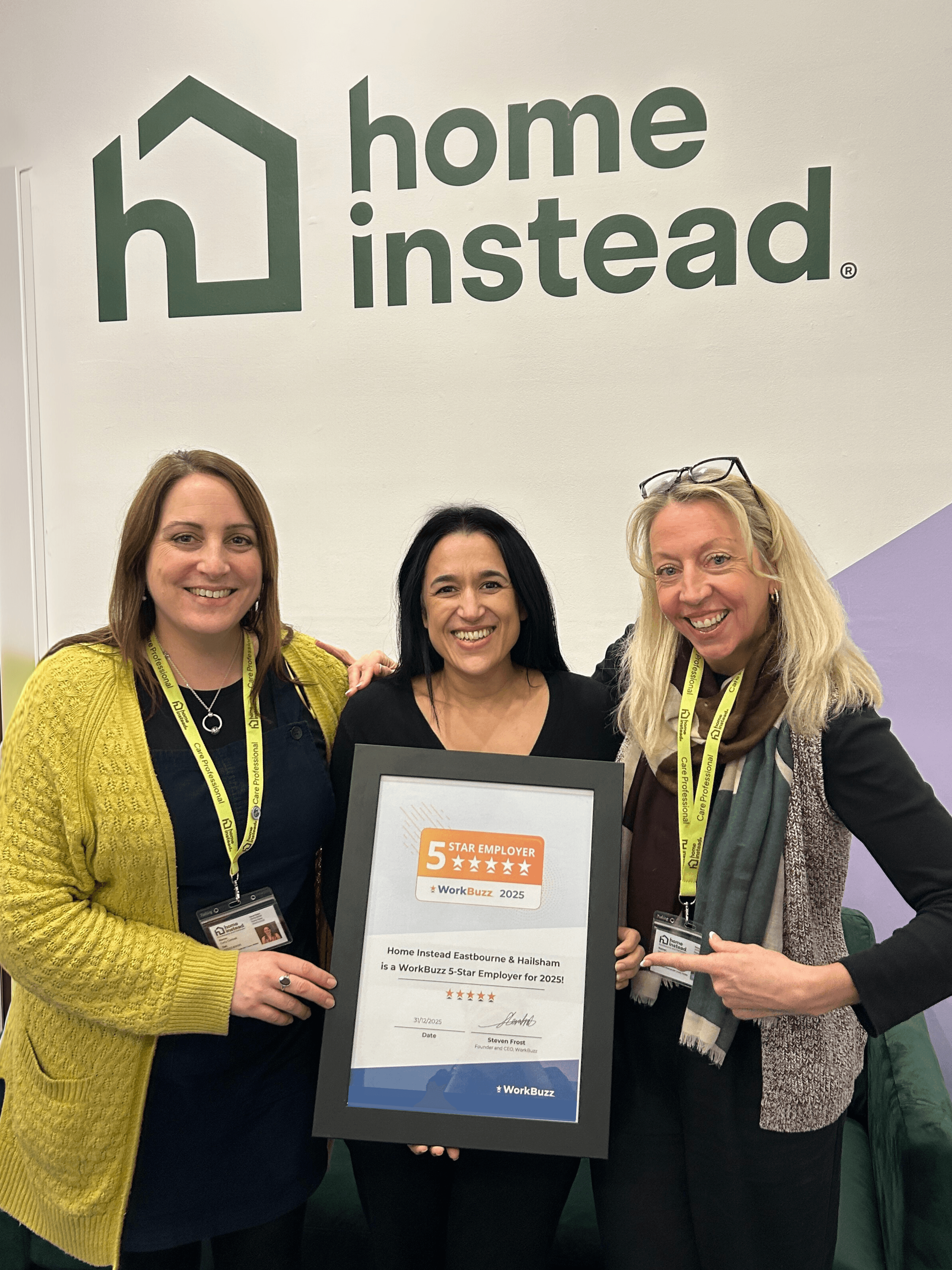 Three smiling women holding a framed award under a "Home Instead" logo on a wall. - Home Instead
