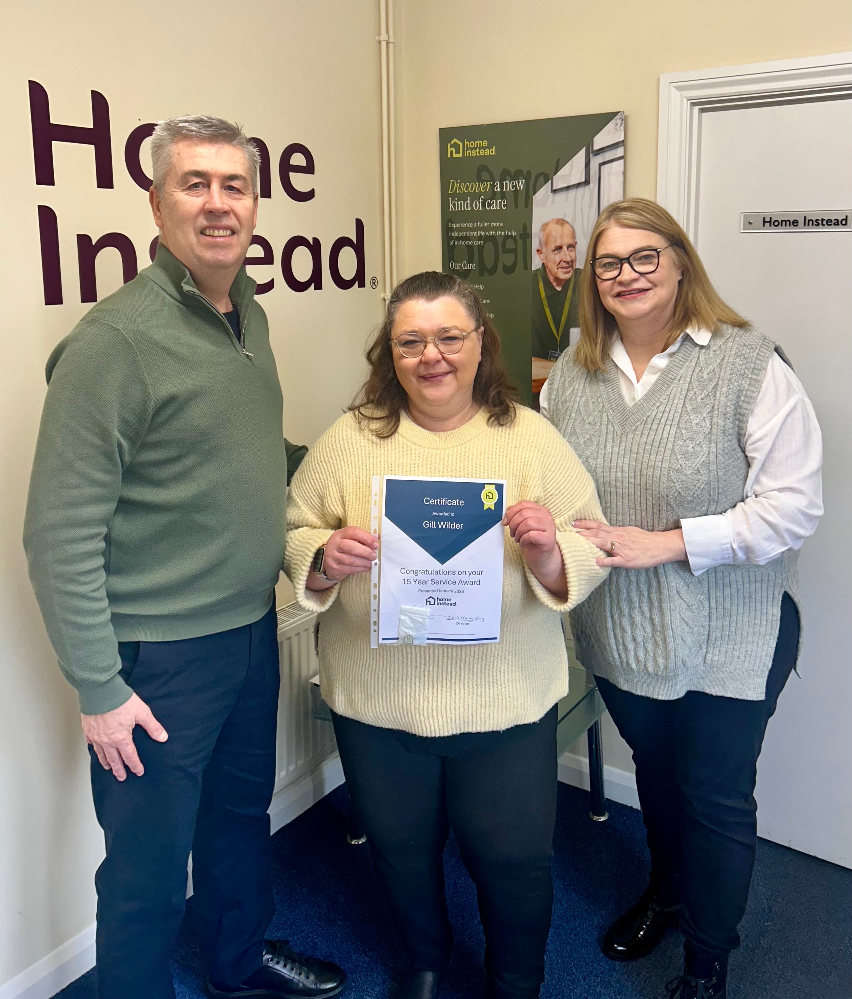 Three people smiling indoors, one holding a certificate, standing in front of a "Home Instead" sign. - Home Instead