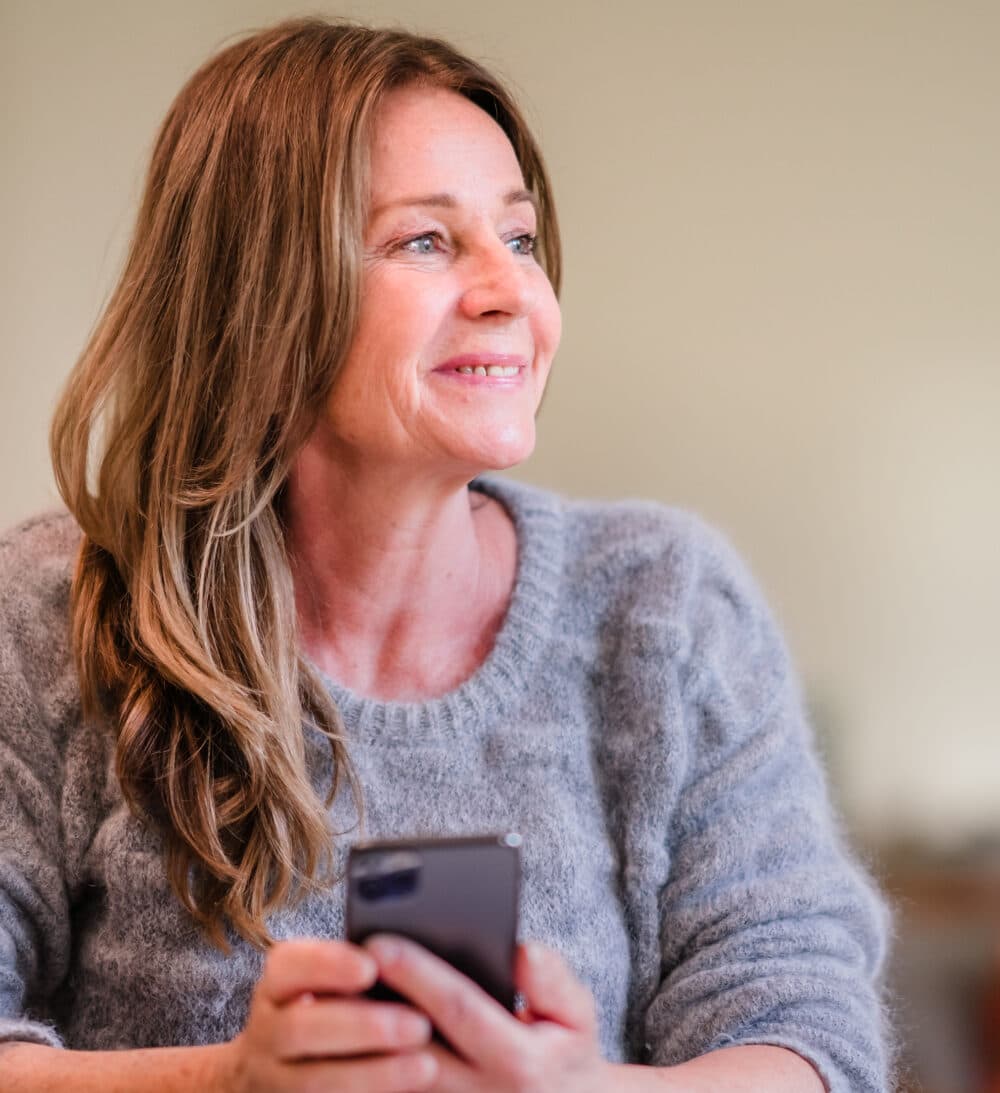 Smiling woman with long hair holding a mobile phone, looking to the side in a cosy indoor setting. - Home Instead