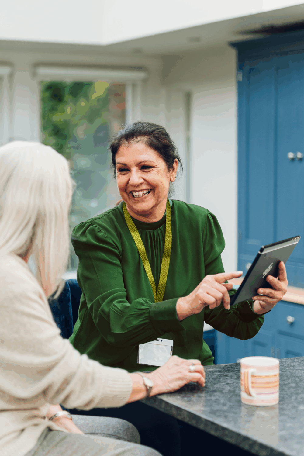 Smiling woman in green showing a tablet to an older woman in a kitchen, both sitting at a worktop. - Home Instead