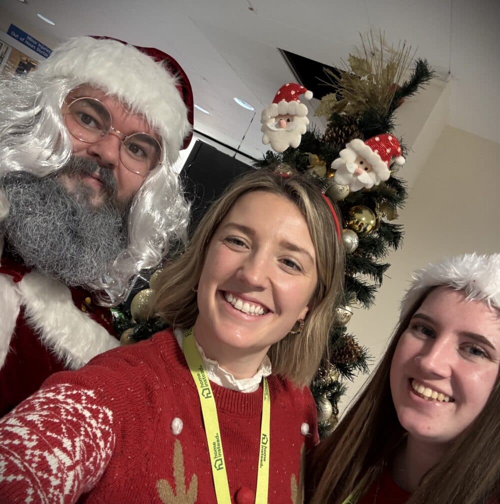 Three people in festive attire smiling in front of a decorated Christmas tree. - Home Instead