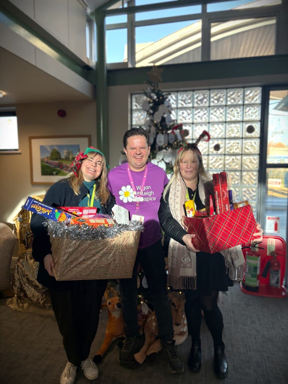 Three people smiling and holding Christmas gift hampers in a festive room with a decorated tree behind them. - Home Instead