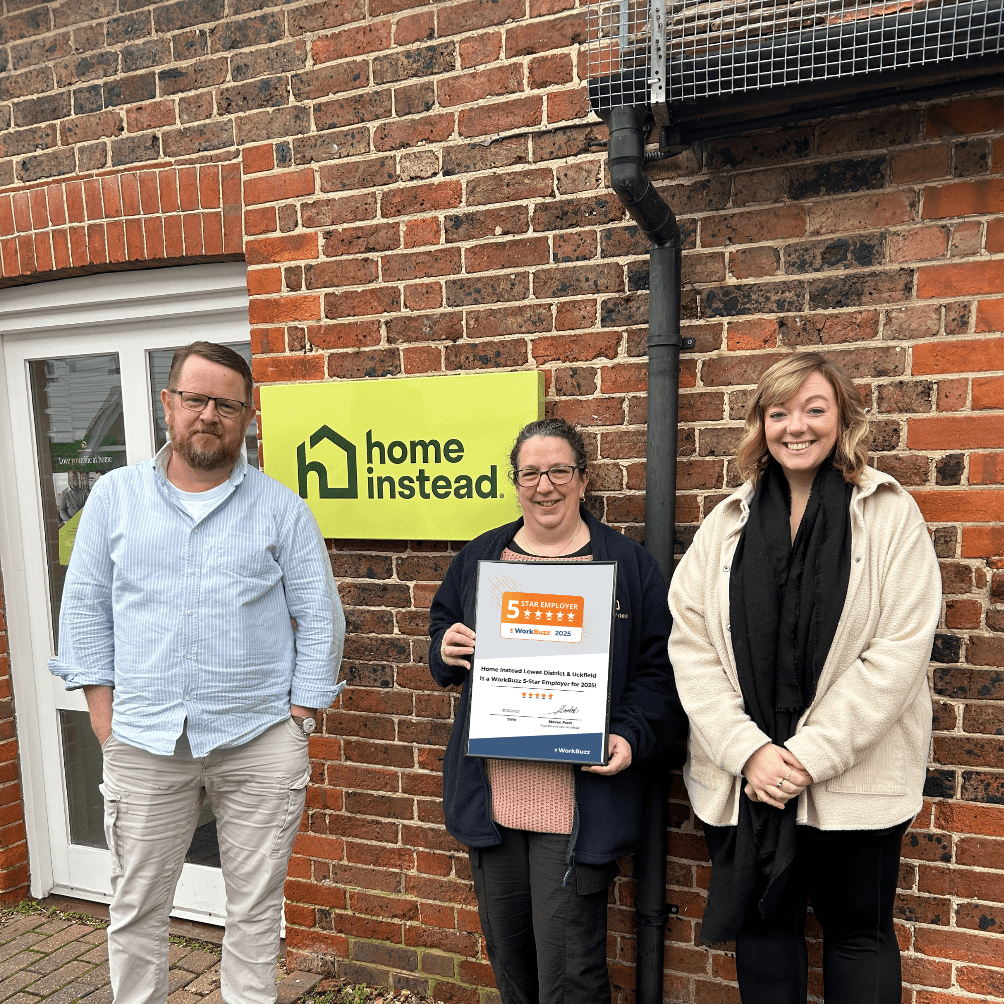 Three Home Instead team members stand outside a brick building, smiling and holding a WorkBuzz 5-Star Employer 2025 certificate.