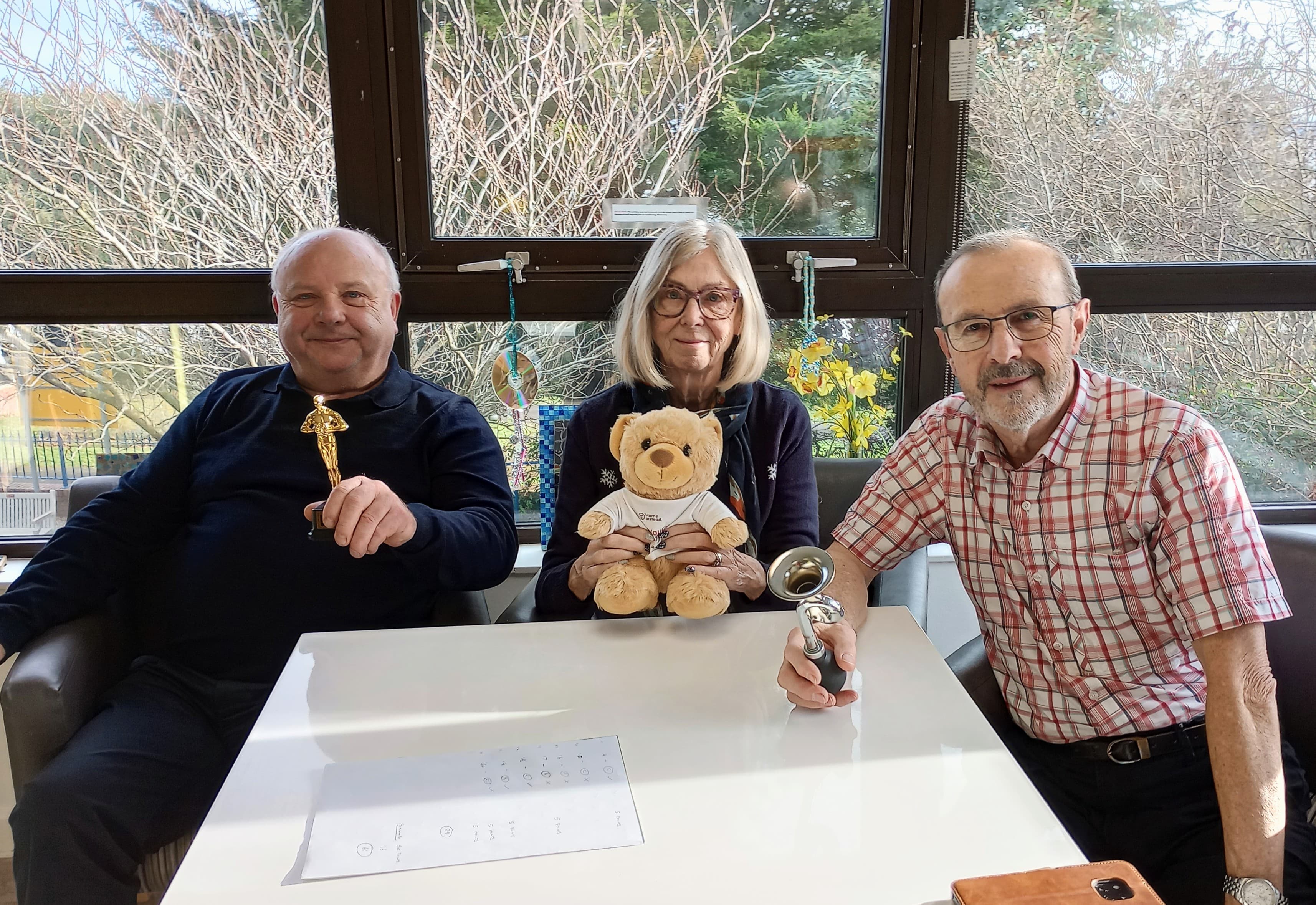 Three older adults sit at a table, holding trophies and a teddy bear, smiling at the camera. - Home Instead