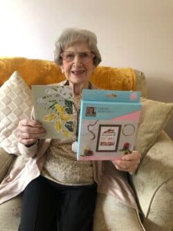 Smiling elderly woman sitting on a sofa, holding a greetings card and a craft kit. - Home Instead