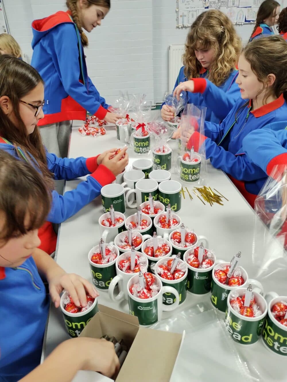 Five girls in blue hoodies fill mugs with chocolates and wrap them in clear plastic at a table. - Home Instead