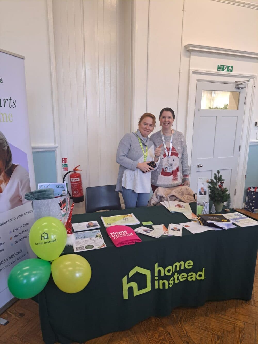 Two women stand behind a Home Instead table with brochures, balloons, and a small Christmas tree on display. - Home Instead
