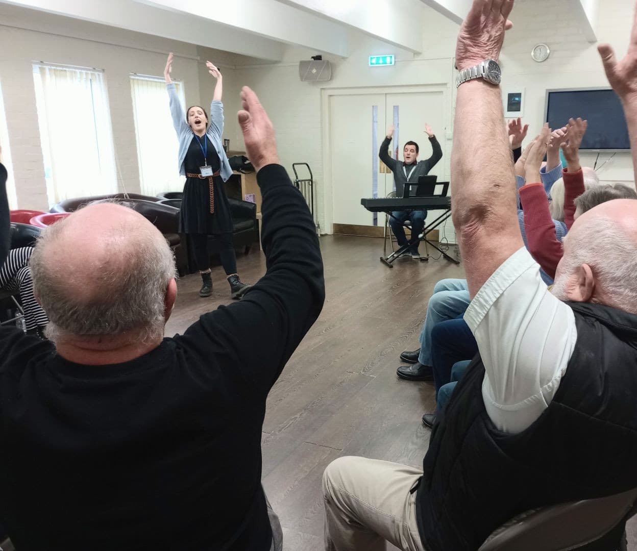 A group of people sit raising their arms as a woman leads and a man plays a keyboard in a bright room. - Home Instead