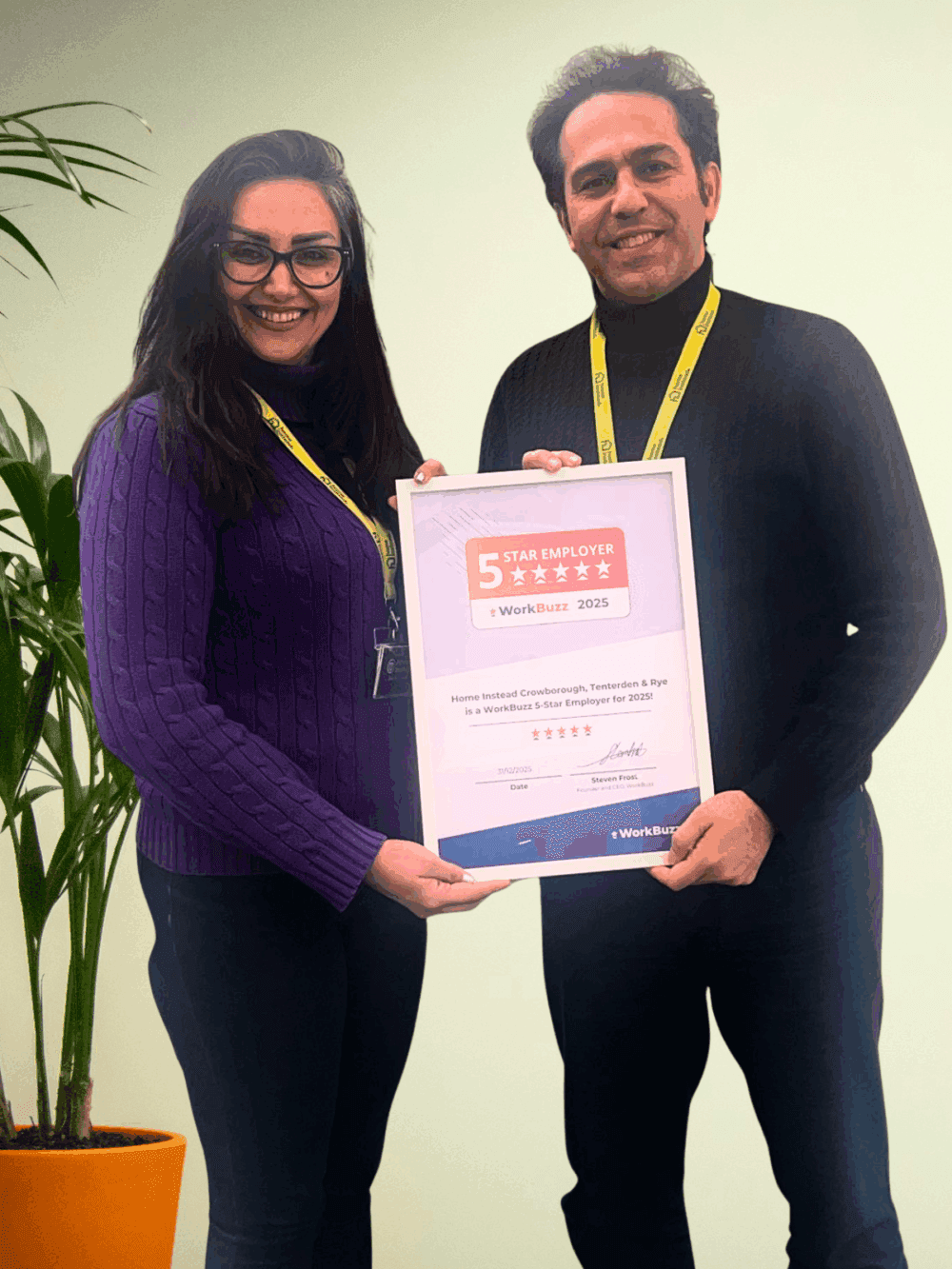 Two people smiling and holding a 5-star employer award, standing beside a green potted plant. - Home Instead