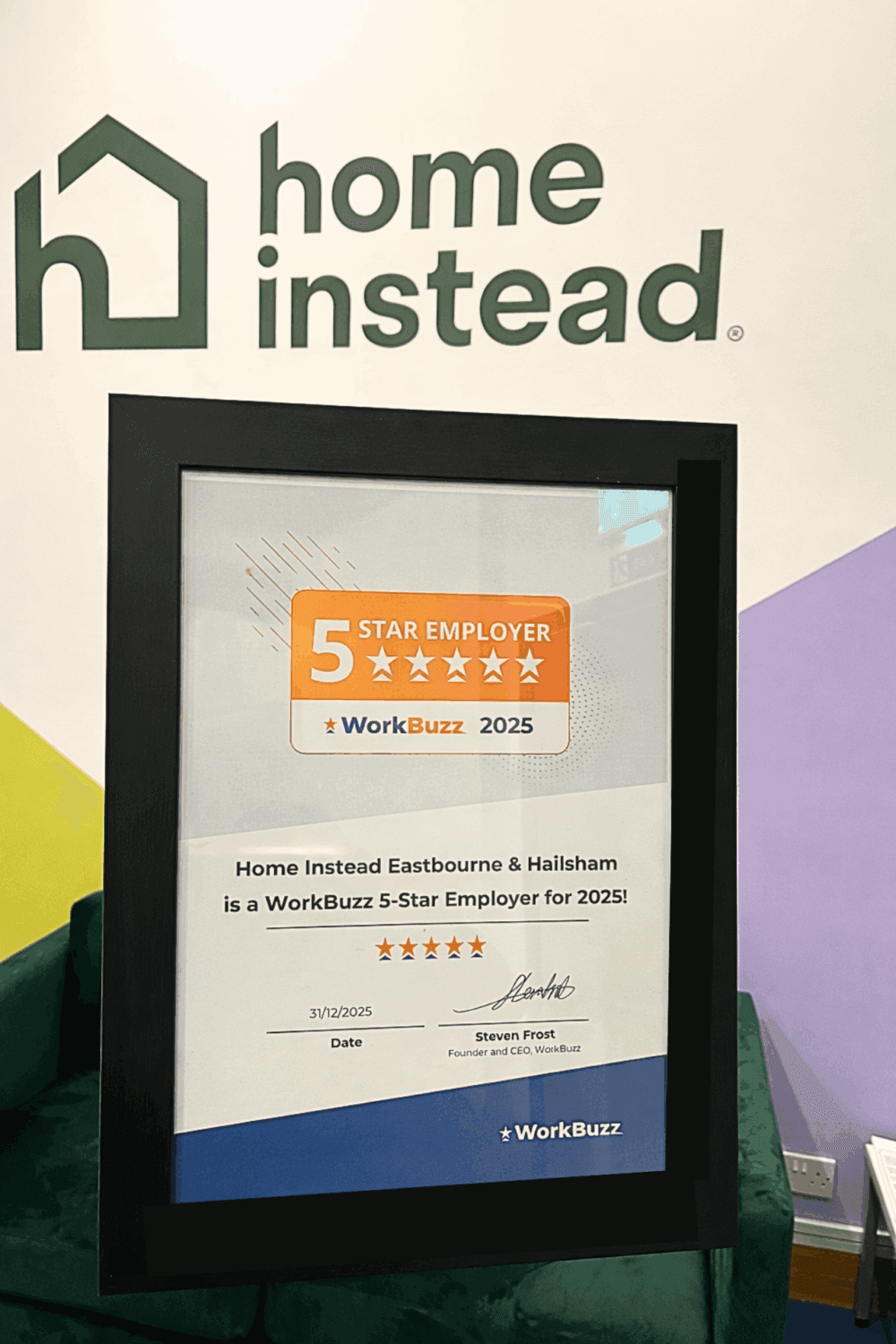 A framed 5-Star Employer 2025 award for Home Instead, displayed in front of a Home Instead logo sign. - Home Instead