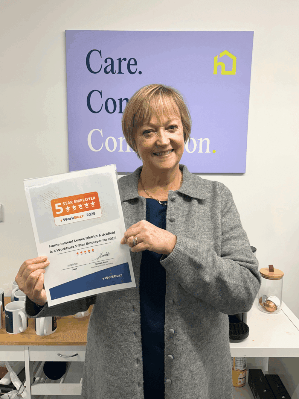 A smiling woman stands indoors holding a framed WorkBuzz 5-Star Employer 2025 certificate, with a Home Instead branded sign on the wall behind her.