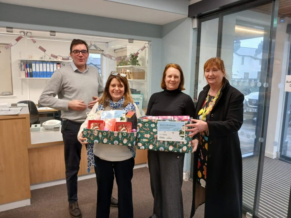 Four people smiling indoors; two hold a festive box filled with food and gift items near a reception desk. - Home Instead