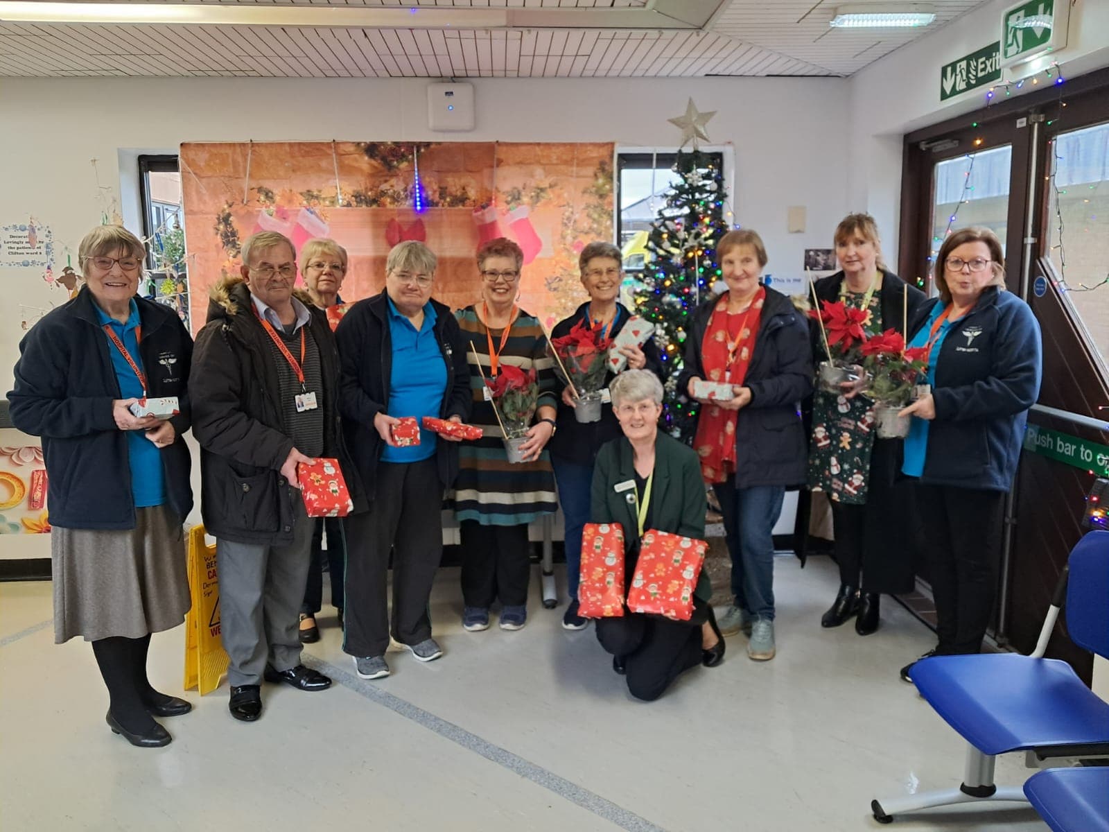 A group of people pose indoors with presents and flowers by a Christmas tree and festive decorations. - Home Instead