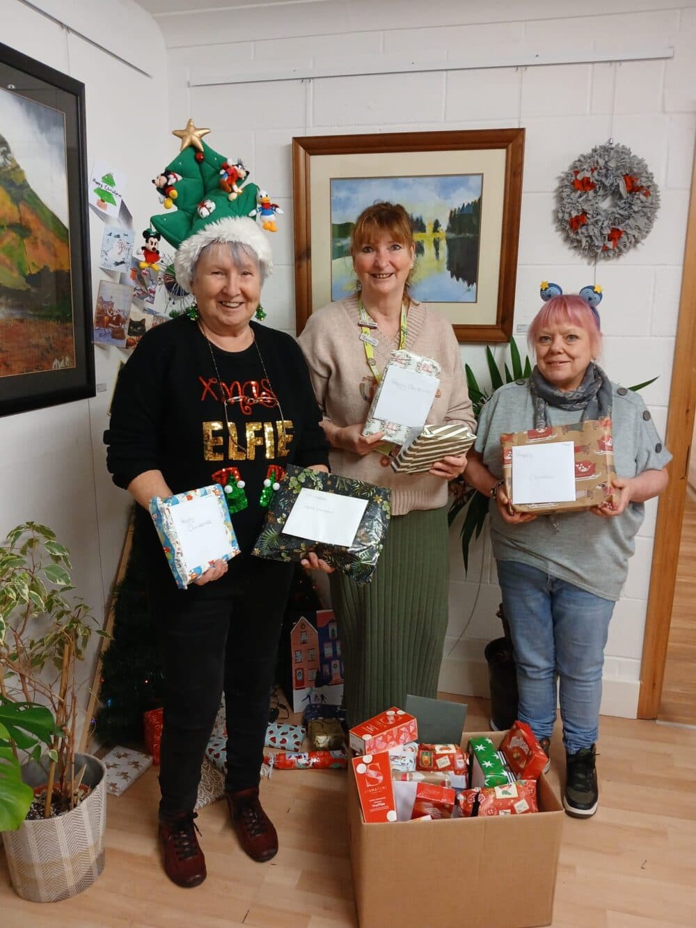 Three smiling women stand by a Christmas tree, holding wrapped gifts and a box of presents. - Home Instead