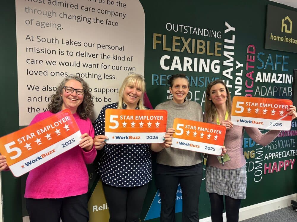Four women smiling and holding "5 Star Employer" signs in front of a wall with positive words and messages. - Home Instead