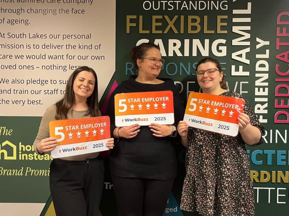 Three women smiling and holding "5 Star Employer 2025" award plaques in front of a colourful wall display. - Home Instead