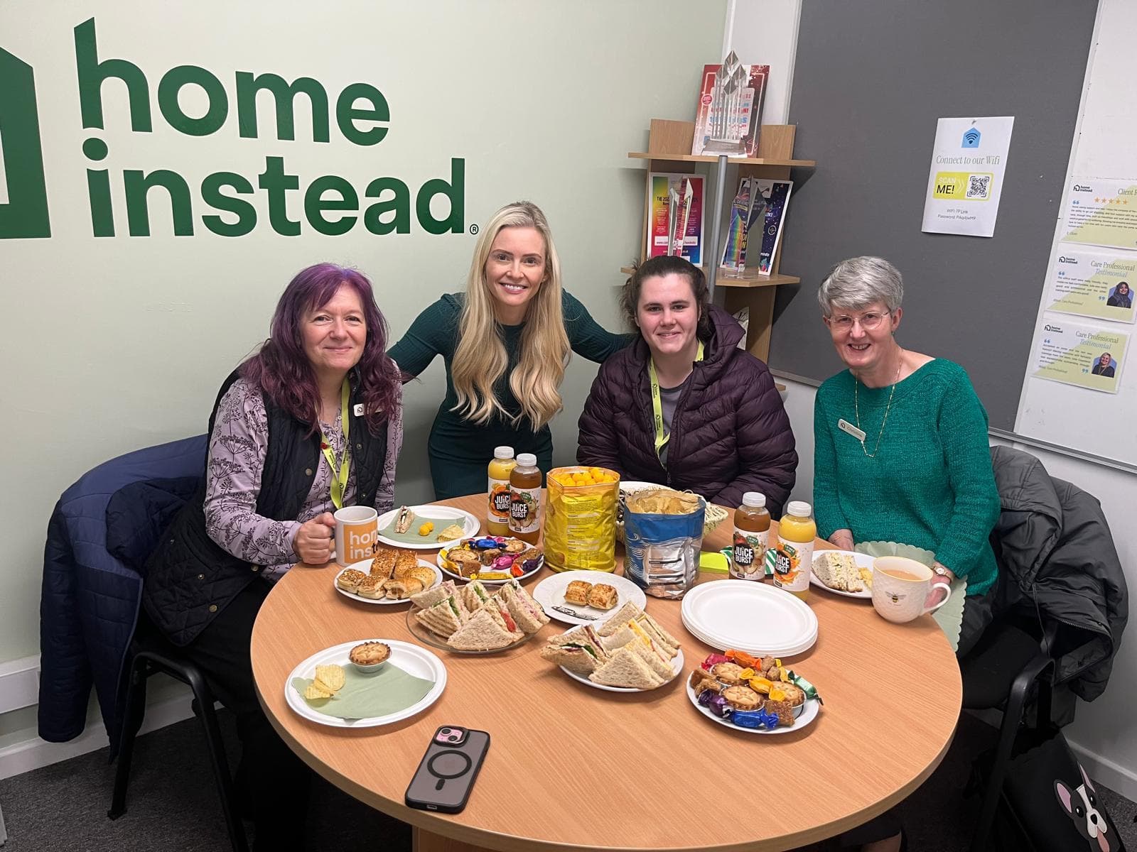Four women sit around a table with snacks and drinks, smiling, in an office with a "home instead" sign. - Home Instead