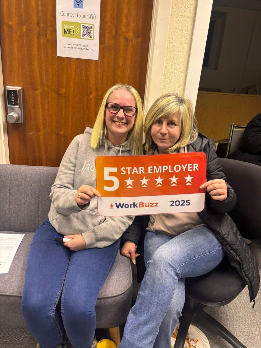 Two smiling women sit on a sofa holding a "5 Star Employer WorkBuzz 2025" sign. - Home Instead