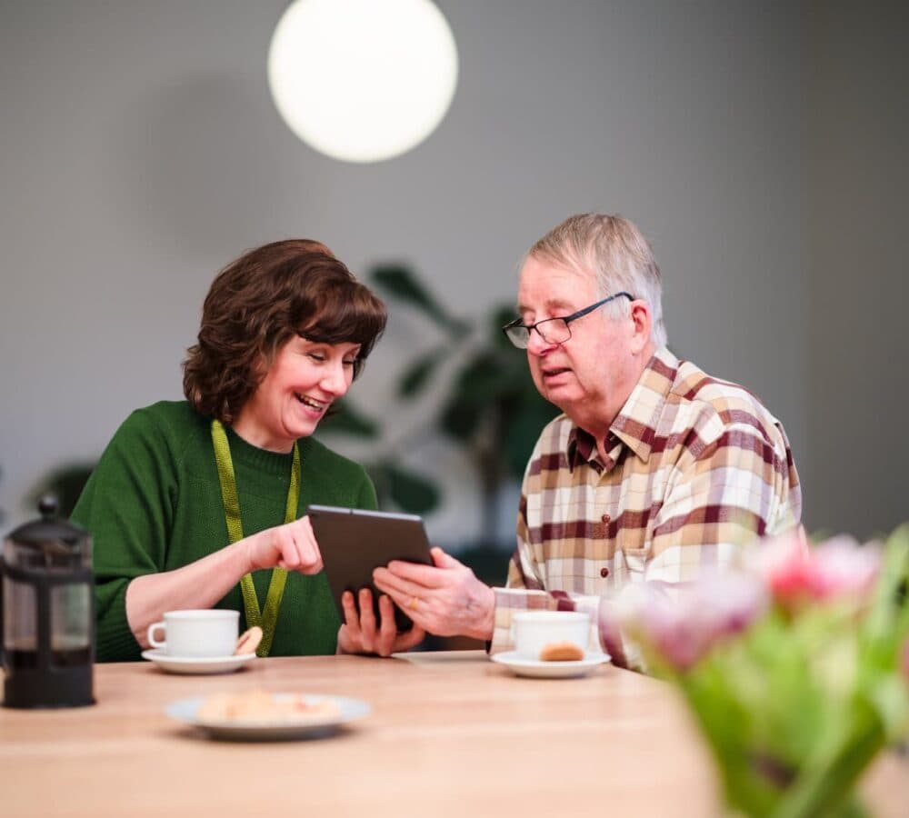 Smiling woman showing an older man something on a tablet at a table with coffee and pastries. - Home Instead