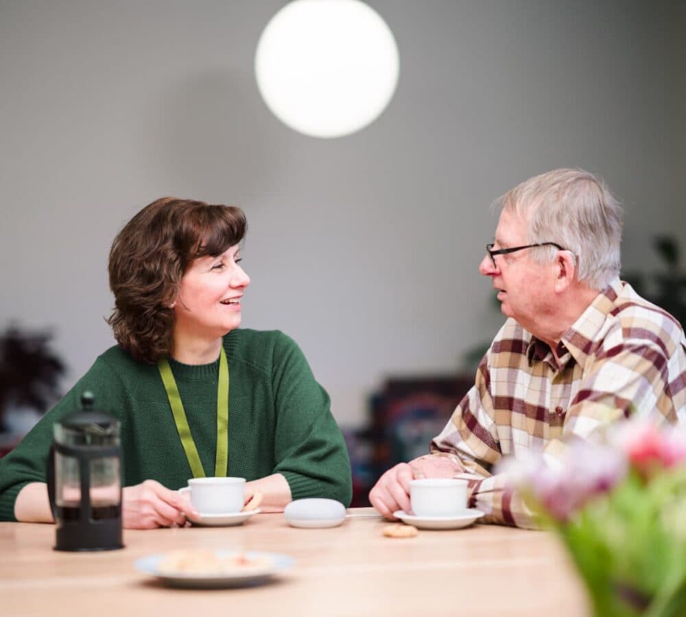 An older male adult with grey hair having a cup of coffee while chatting with his younger female carer