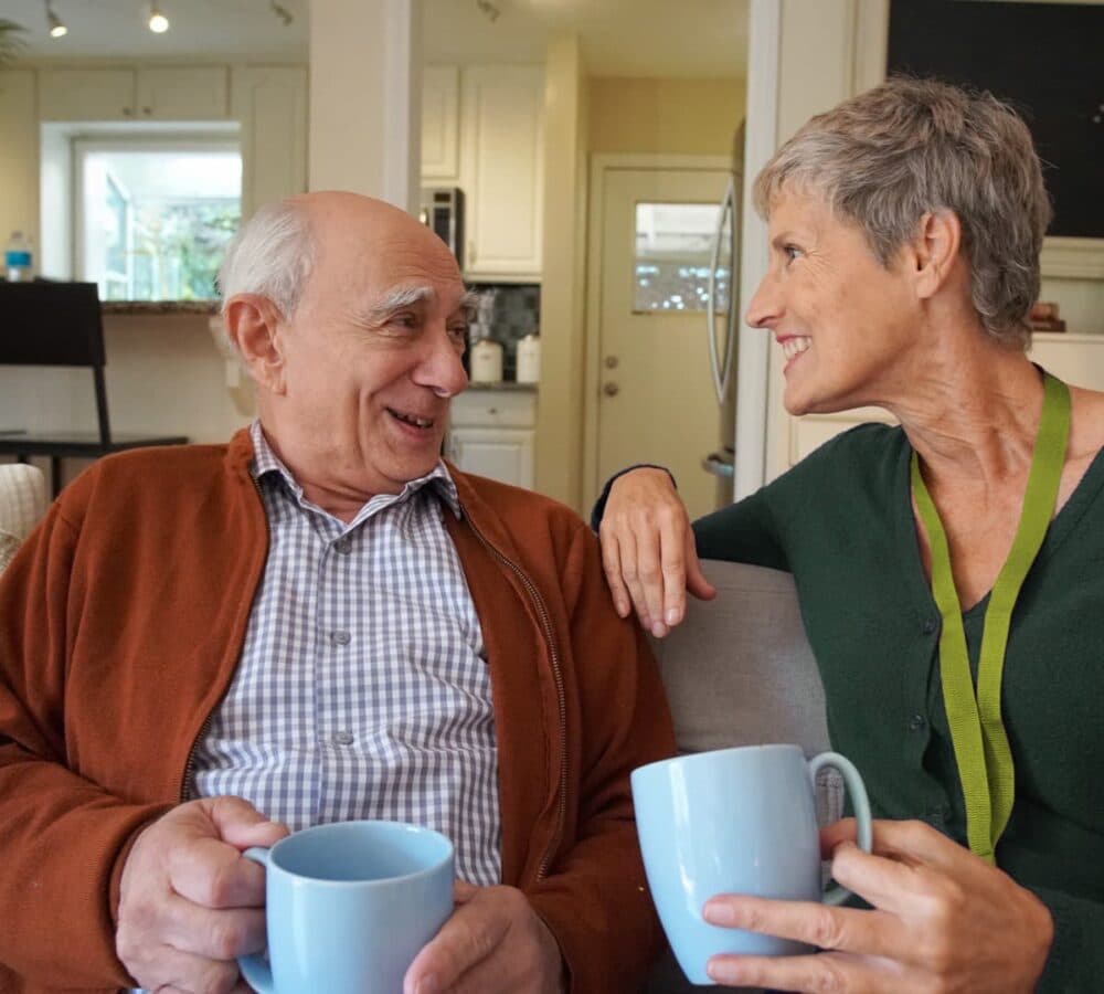 An older male adult with grey hair having coffee and happy and smiling while sitting on the couch with his younger female carer inside the house