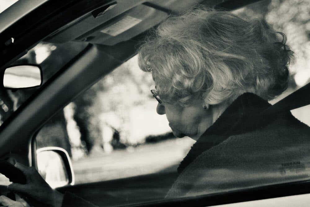 Elderly woman with glasses driving a car, viewed from the passenger side window. - Home Instead