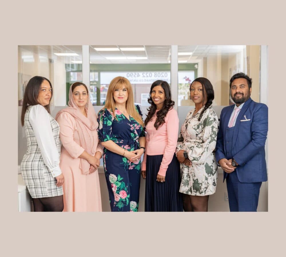 Six professionally dressed people standing in a row, smiling, in an office setting. - Home Instead