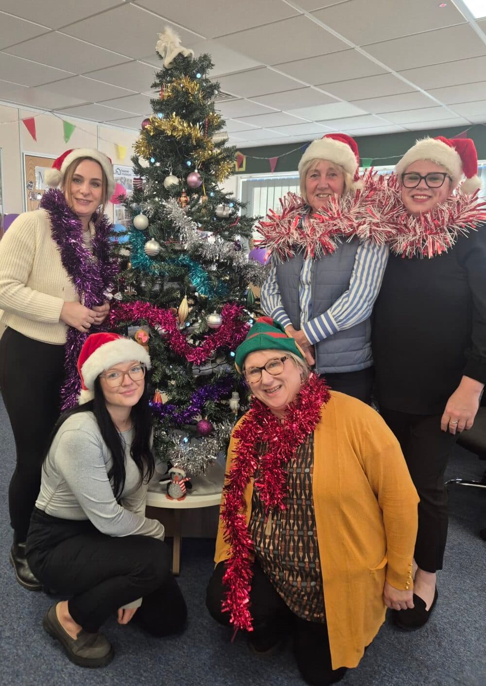 Five women in festive hats and tinsel pose by a decorated Christmas tree in an office setting. - Home Instead