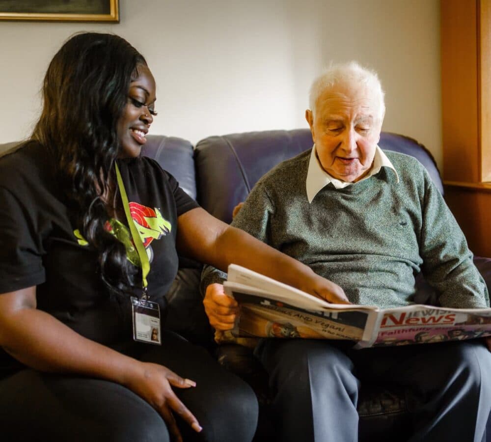 An older male adult looking at the newspaper with his female carer with black hair both sitting on the couch