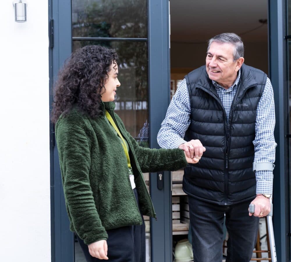 An older male adult with grey hair and using a crutch going out of the door with his younger female carer with long black curly hair