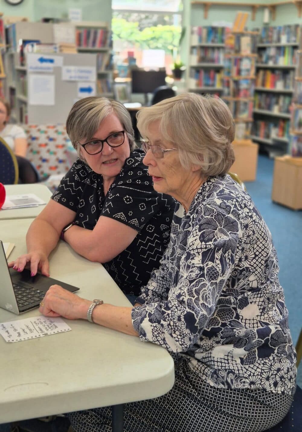 Two older women sit at a table in a library, looking at a laptop together and talking. - Home Instead