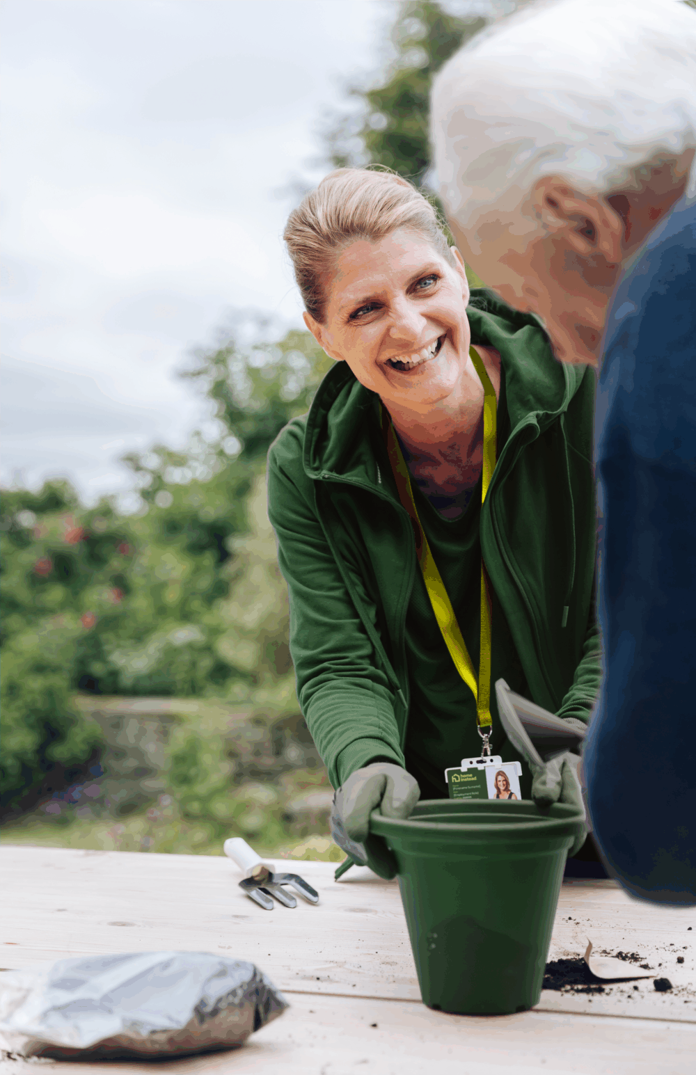 A smiling carer helps an elderly person with gardening, holding a green flowerpot outdoors. - Home Instead