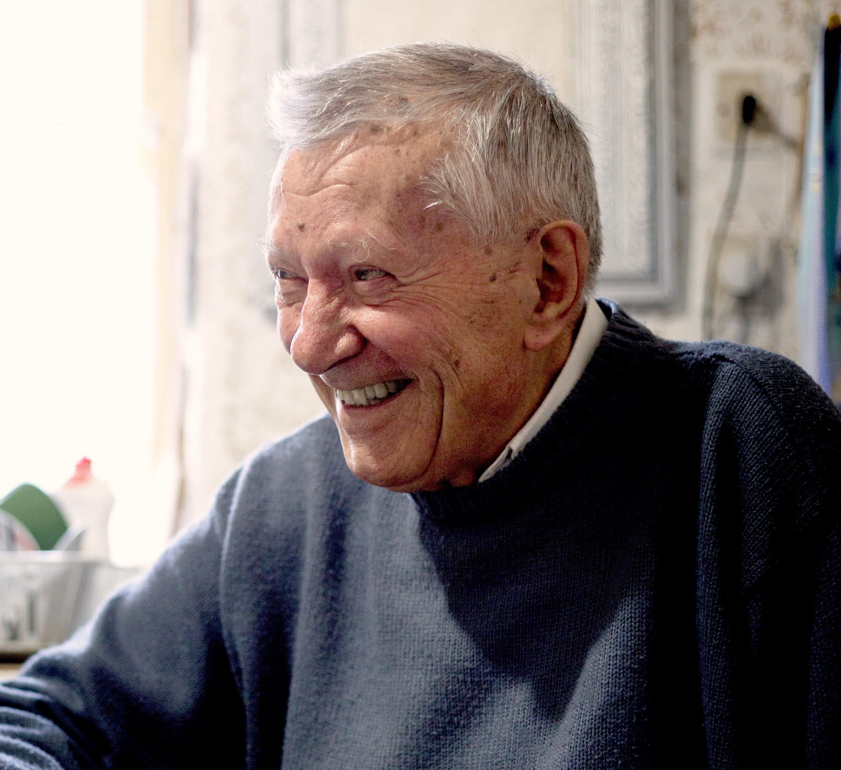 Elderly man sitting at his kitchen table, laughing, with a walking stick by his side