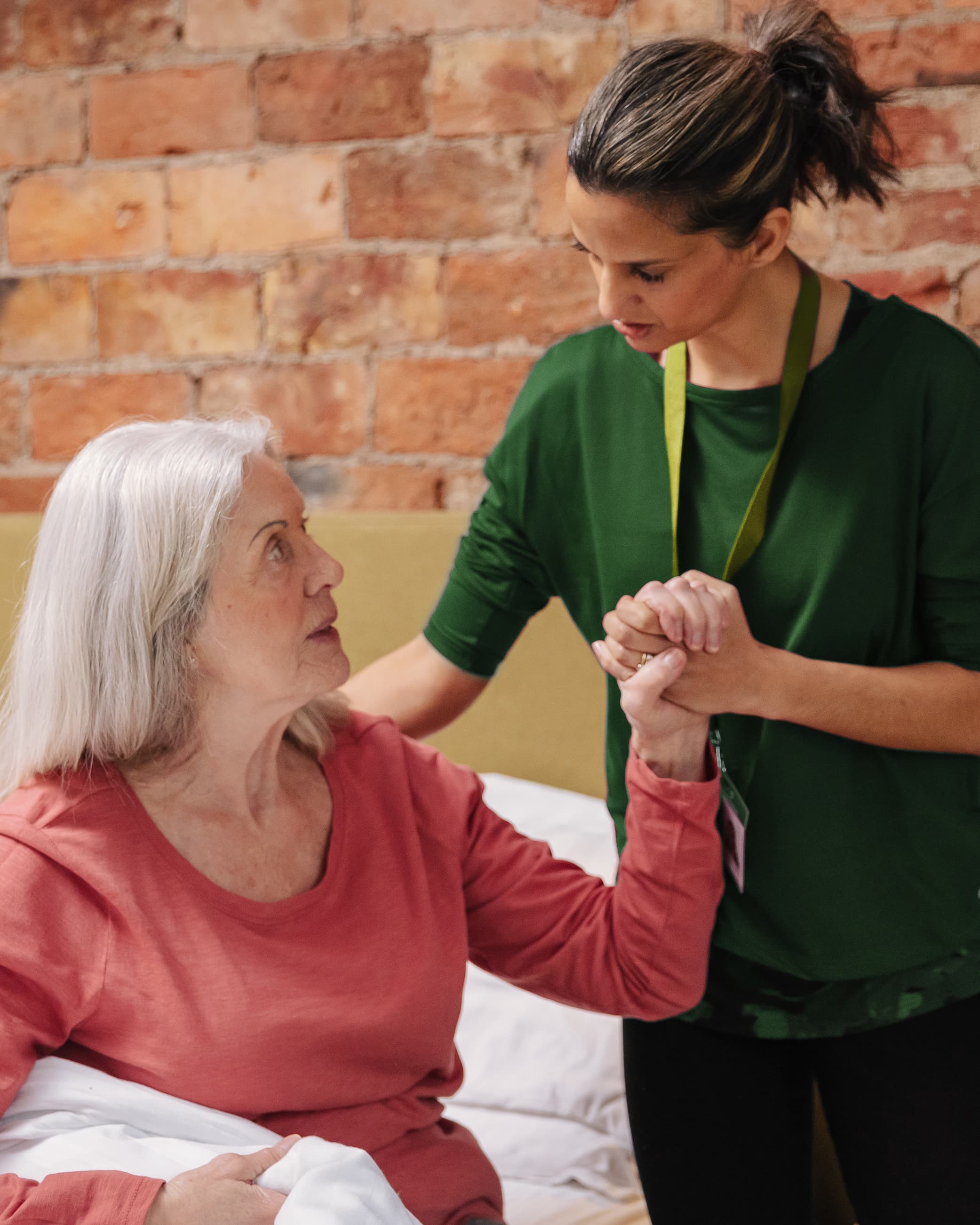 A carer supports an elderly woman sitting on a bed, holding her hand and offering comfort. - Home Instead