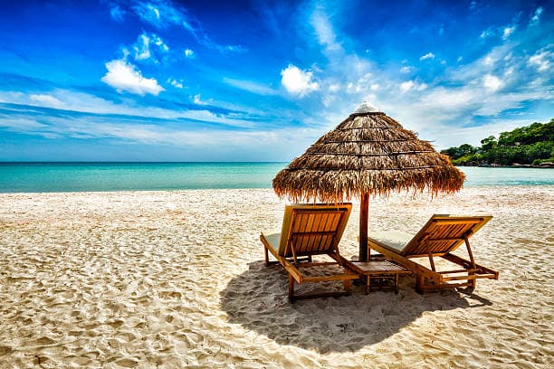 Two wooden sun loungers under a thatched parasol on a sandy beach with a blue sky and calm sea. - Home Instead