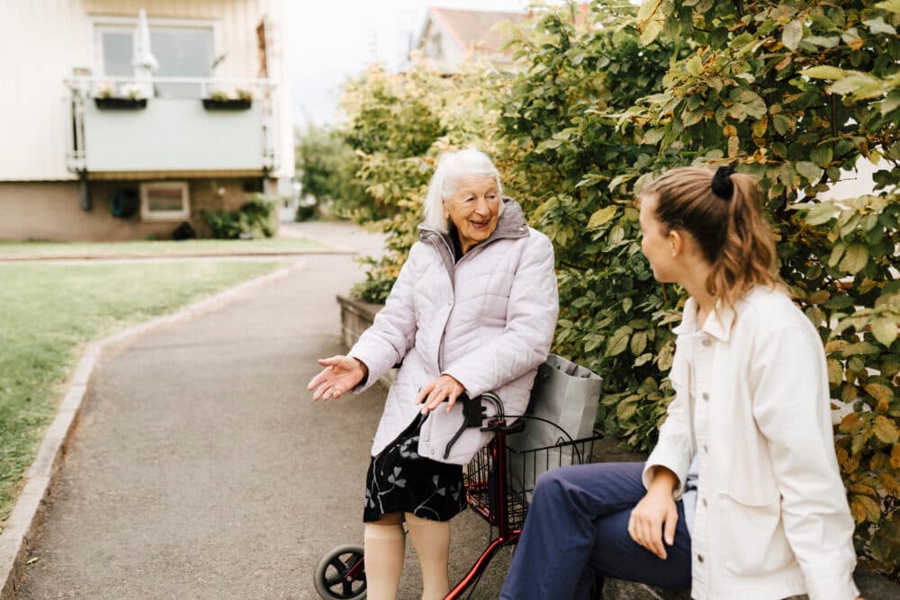 Elderly woman on a zimmer frame talking with a young woman on a pavement near bushes outside. - Home Instead