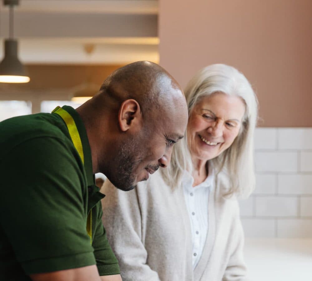 An older female adult with long white hair happy and smiling while preparing food with her male carer wearing green