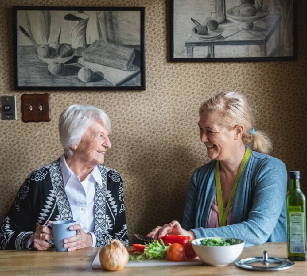 An older female adult with short grey hair smiling while drinking coffee with her younger female carer chopping vegetables inside the kitchen