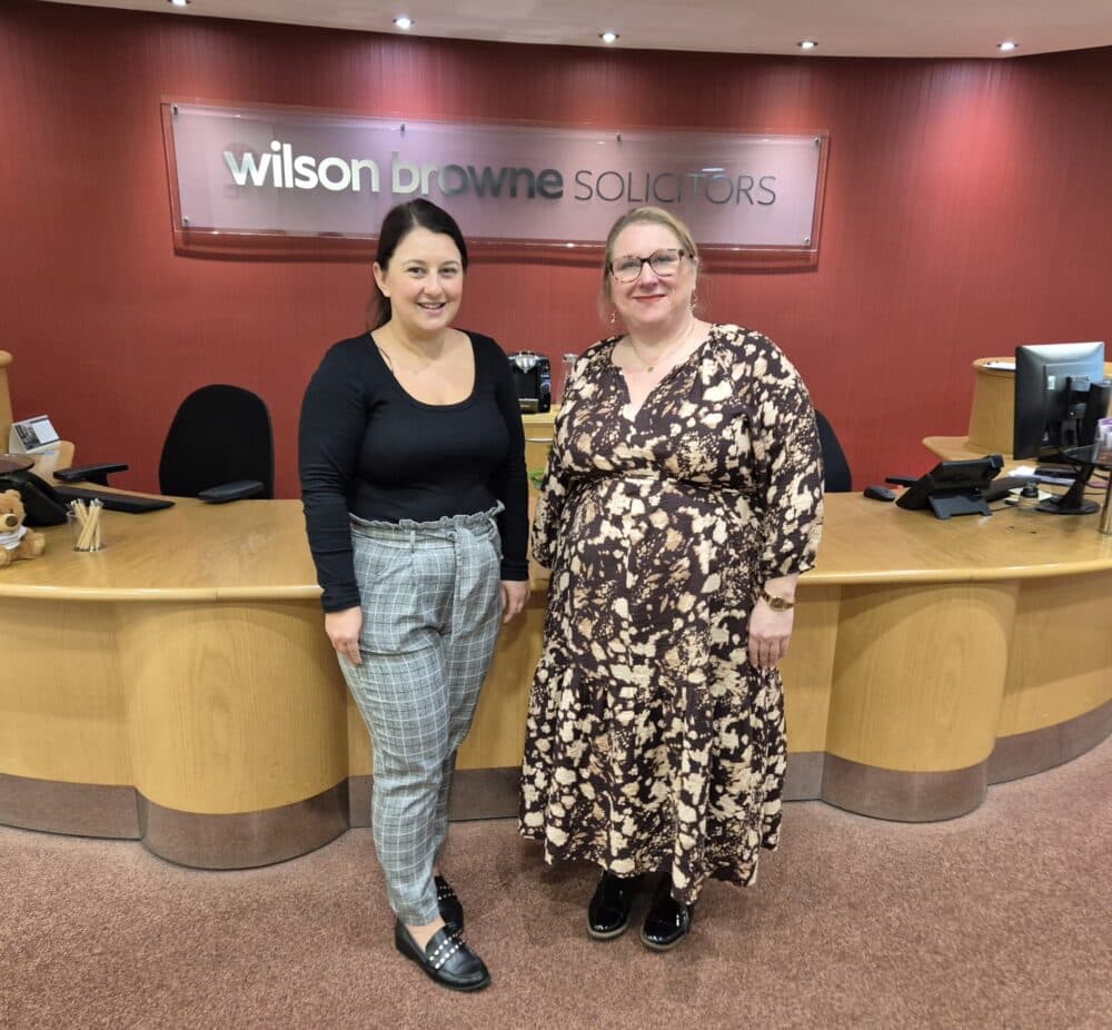 Two women standing and smiling in front of a reception desk at Wilson Browne Solicitors office. - Home Instead