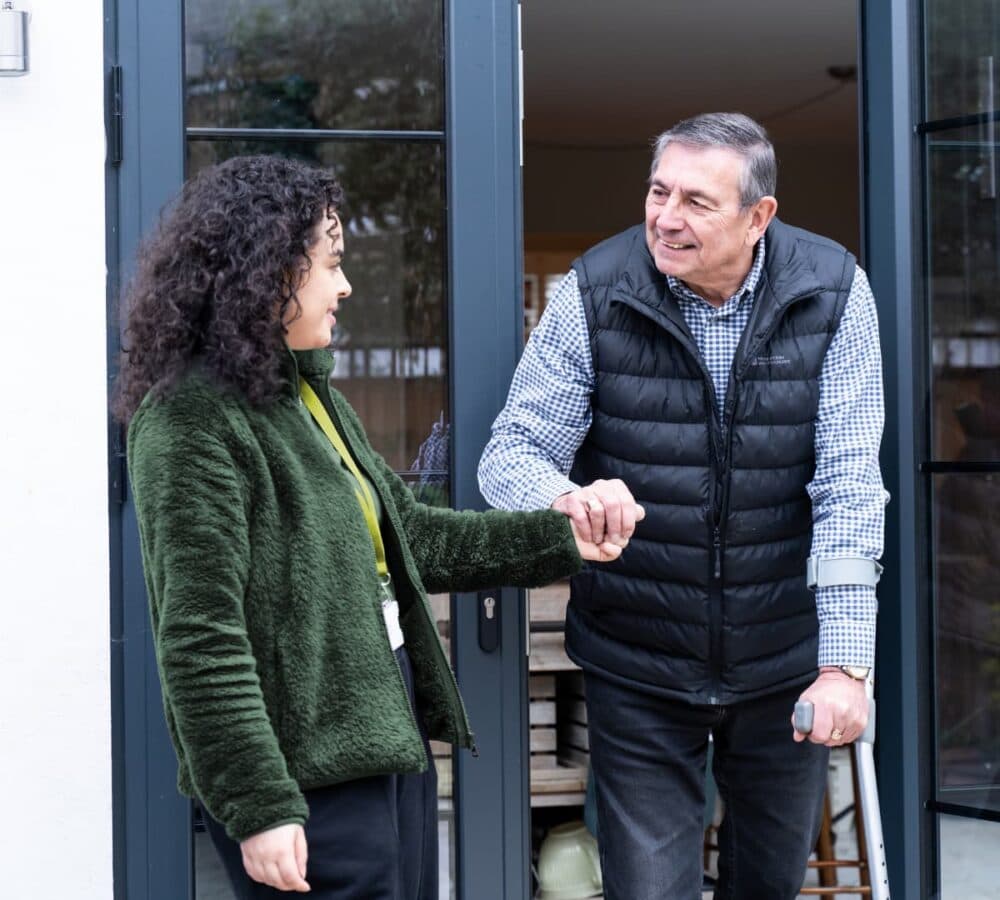 An older male adult with grey hair smiling and wearing a puffer jacket and using a crutch while walking out of the house and being helped by his younger female carer with curly hair