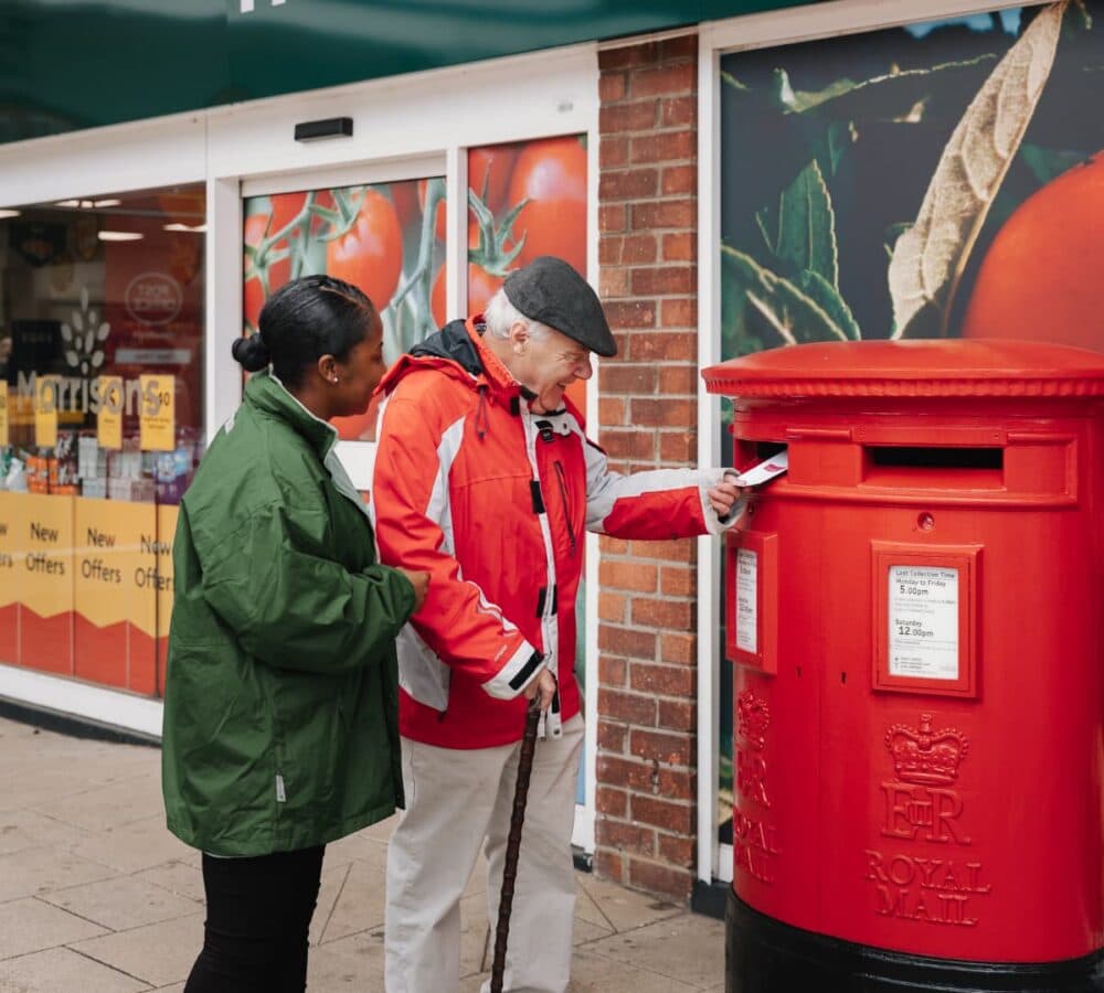 An older male adult mailing a letter and using a crane with his younger female carer wearing green jacket
