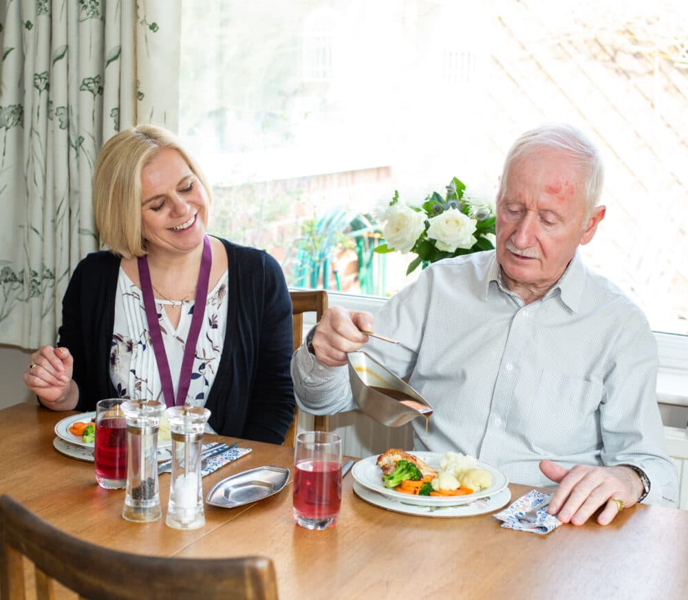 Elderly man and young female carer sat at the dinner table together, and the man is pouring gravy onto his dinner
