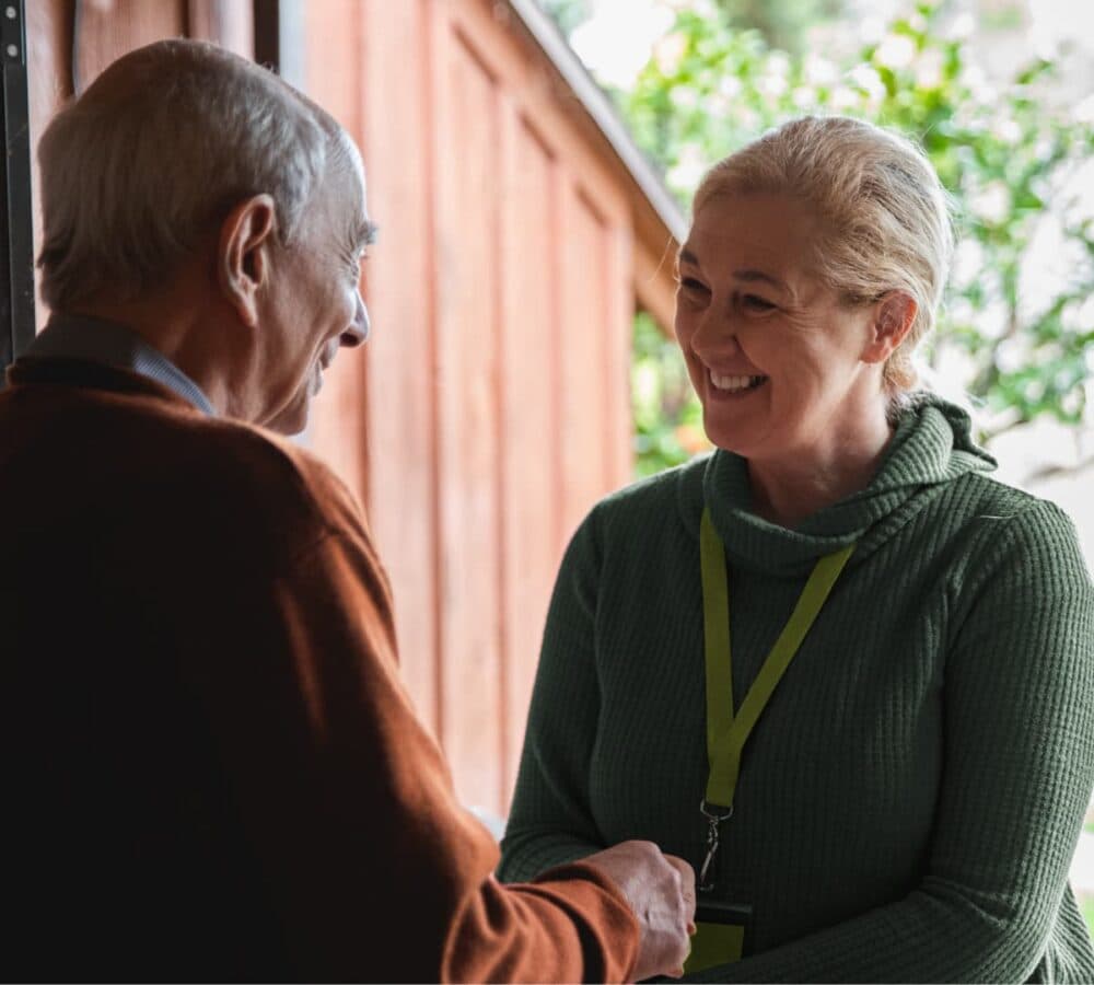 An older male adult with grey hair greeting a woman with blonde hair and wearing green shaking her hands at the door