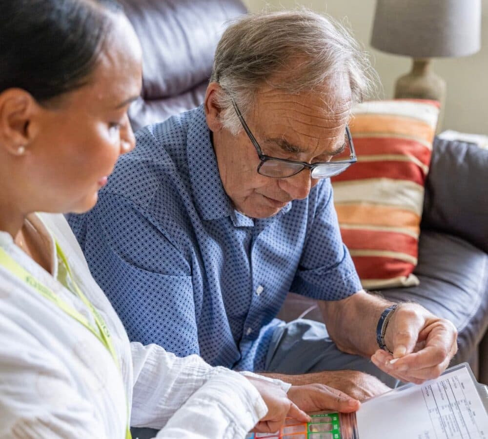 An older male adult with grey hair wearing eyeglasses and taking medicine with his younger femaile carer helping her while sititing on a couch