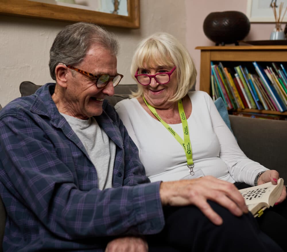 Female Home Instead live-in carer sitting in lounge with her elderly male client looking at a book of crossword puzzles together