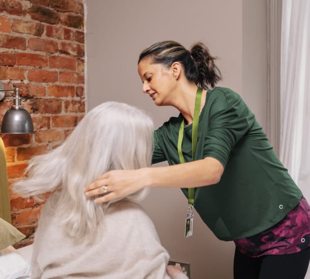 An older female adult with long white hair sitting on a bed while her hair is being combed by her younger female carer wearing green